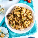 A white bowl filled with grilled, seasoned chicken pieces garnished with fresh herbs, placed on a blue napkin with cherry tomatoes and skewers nearby.