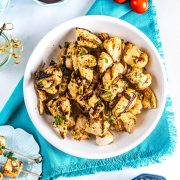 A white bowl filled with grilled, seasoned chicken pieces garnished with fresh herbs, placed on a blue napkin with cherry tomatoes and skewers nearby.