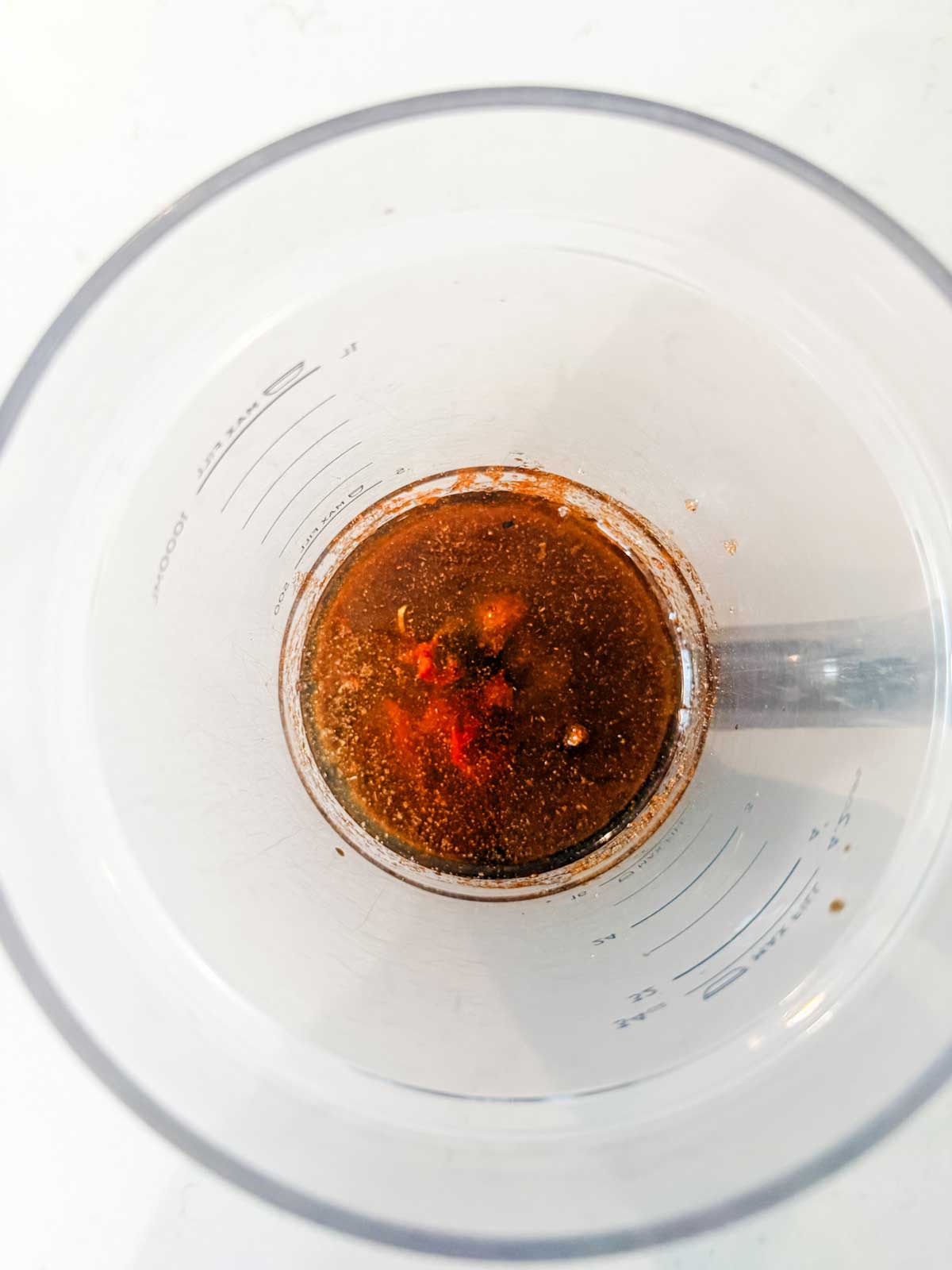 A top-down view of a measuring cup containing a brown liquid mixture with visible spices and small chunks, placed on a white surface.