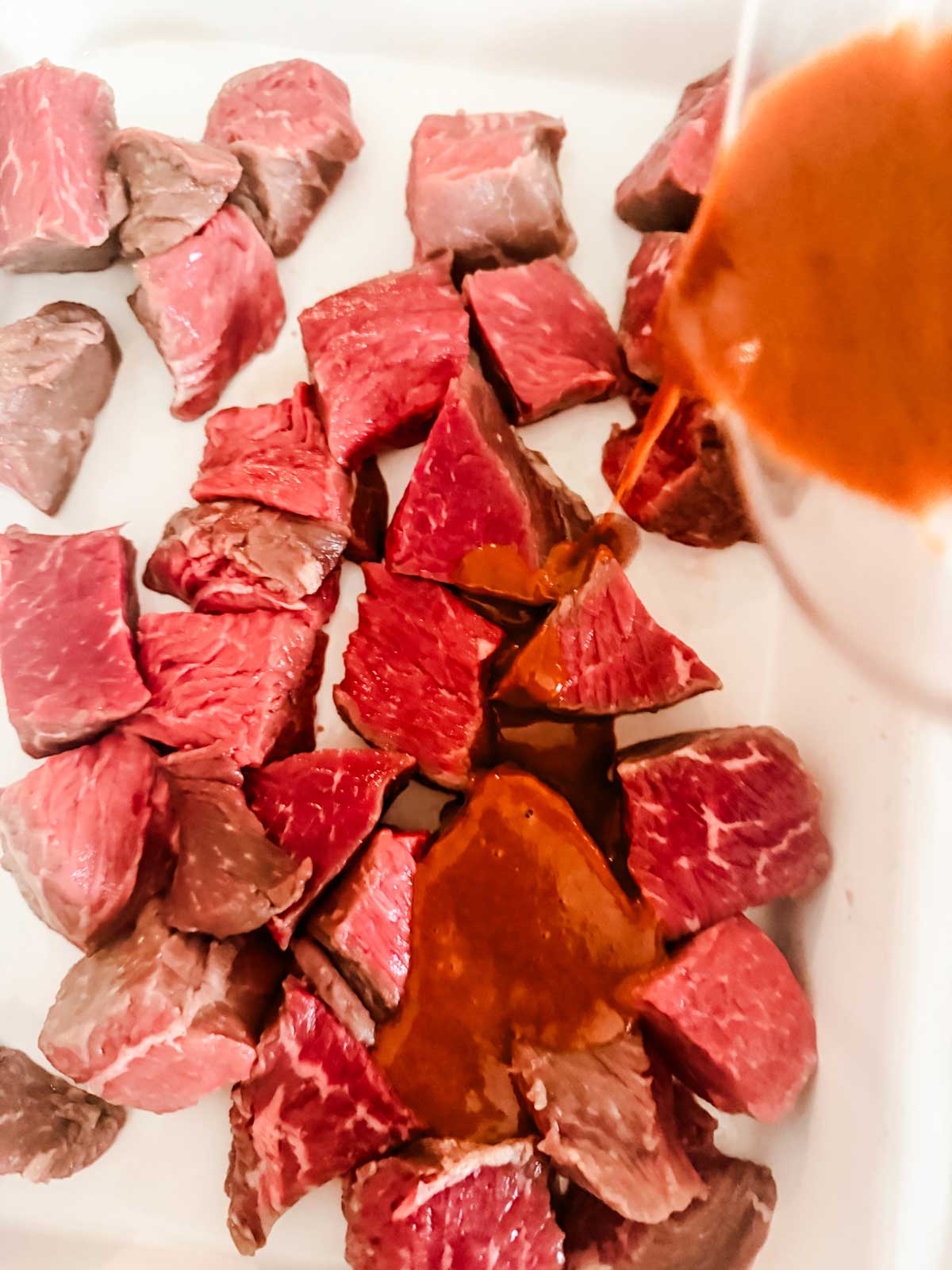 Raw beef chunks placed in a dish with a reddish-brown marinade being poured over them from a container, preparing the meat for cooking.