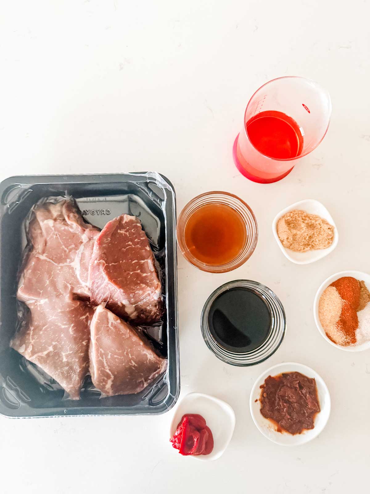 Raw steak in a black plastic tray next to small bowls containing brown sugar, spices, soy sauce, ketchup, and Worcestershire sauce, with a glass of red liquid on a white surface.
