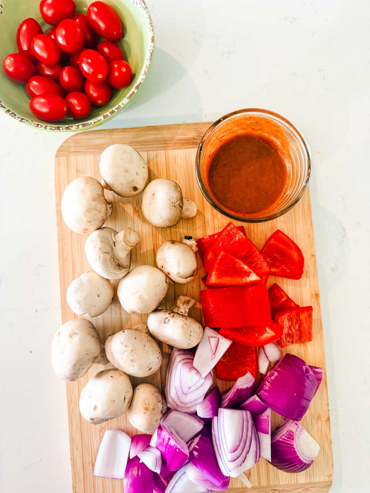 A cutting board with whole white mushrooms, chopped red bell pepper, and chopped red onion, next to a small bowl of reddish sauce. A separate bowl holds grape tomatoes.