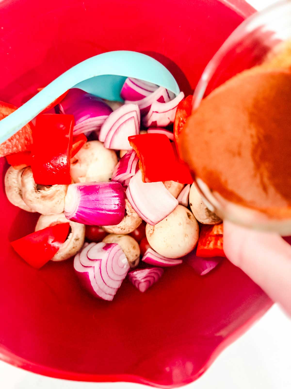 A red mixing bowl filled with chopped red onions, red bell peppers, and whole mushrooms, with a blue spatula and sauce being poured over the vegetables.