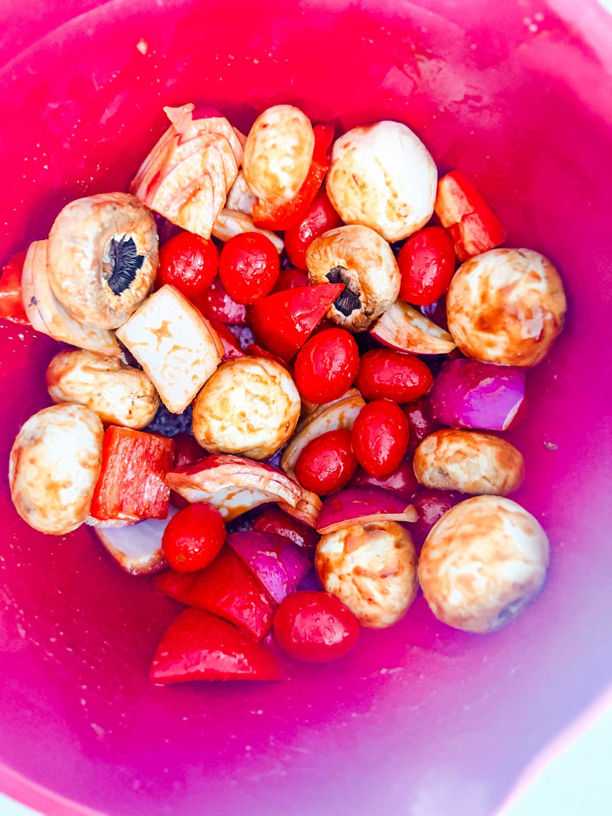 A pink bowl filled with fresh vegetables, including whole mushrooms, cherry tomatoes, chopped red onions, and chunks of red bell pepper, all coated in a marinade.