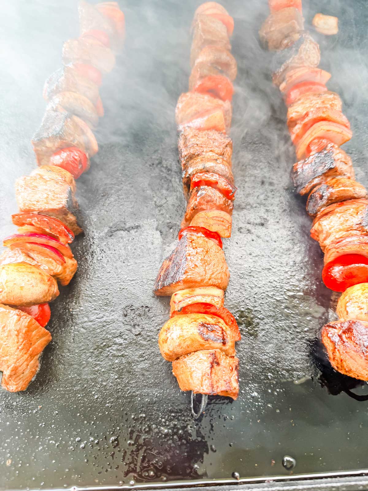 Three skewers with grilled beef, mushrooms, and red bell peppers cooking on a smoky grill.