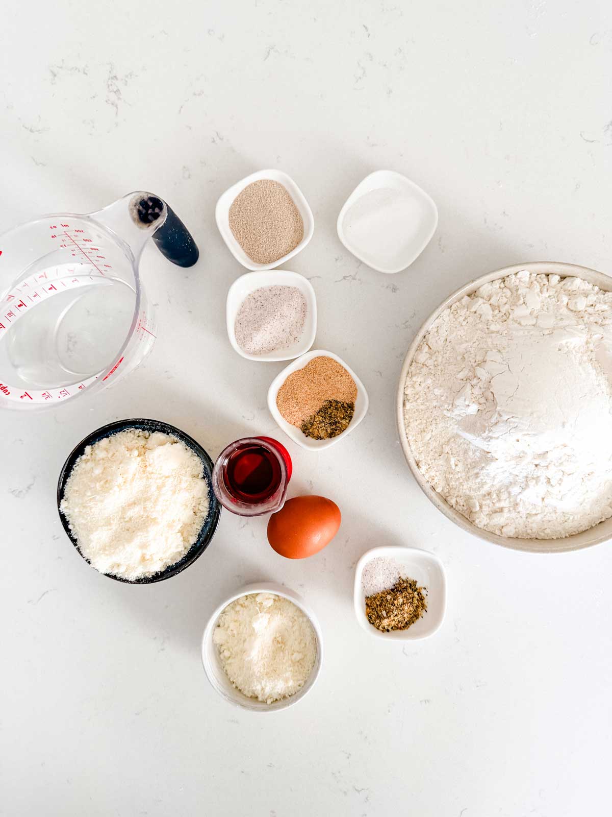 Various baking ingredients are arranged on a white surface, including flour in a bowl, a measuring cup with water, an egg, grated cheese, spices, yeast, sugar, and a small jar of red liquid.