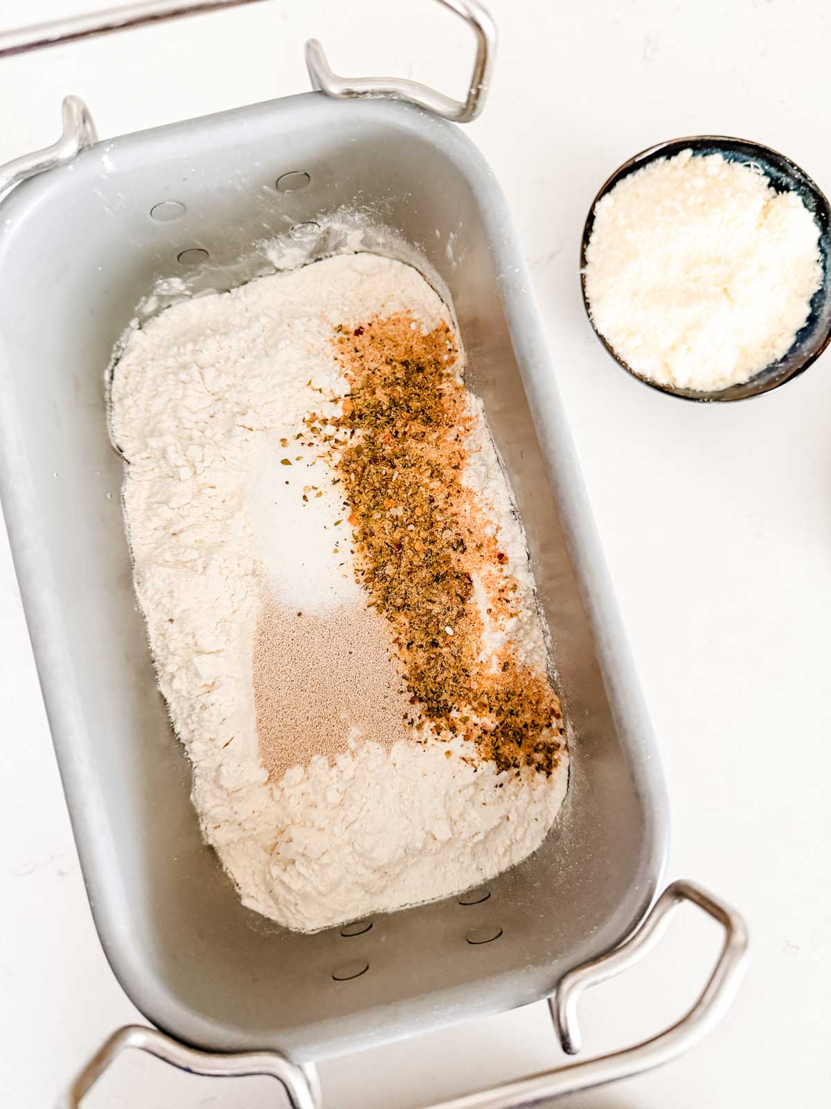 A bread machine pan filled with flour, dry yeast, and seasonings, next to a small bowl of grated cheese on a white surface.