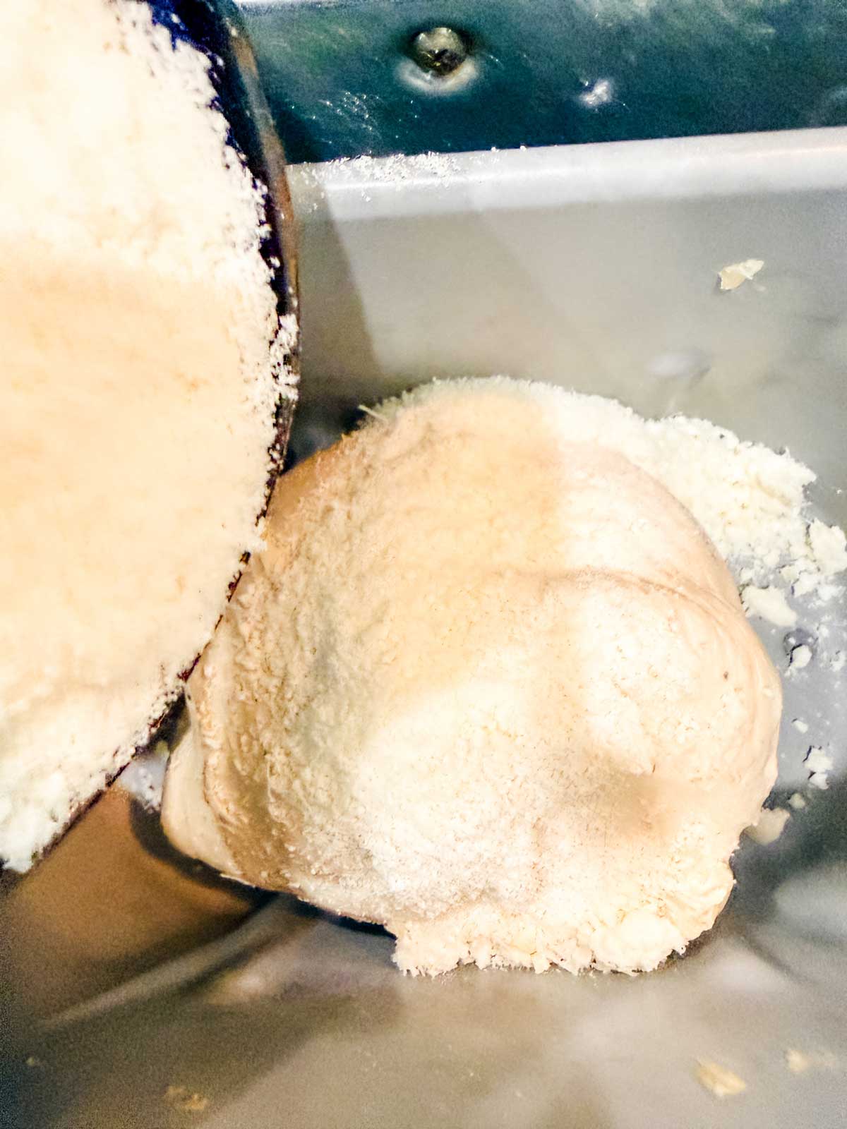 A close-up of pizza dough being kneaded in a stand mixer bowl, with flour dusted on top of the dough. The metal mixing attachment is partially visible on the left side of the image.