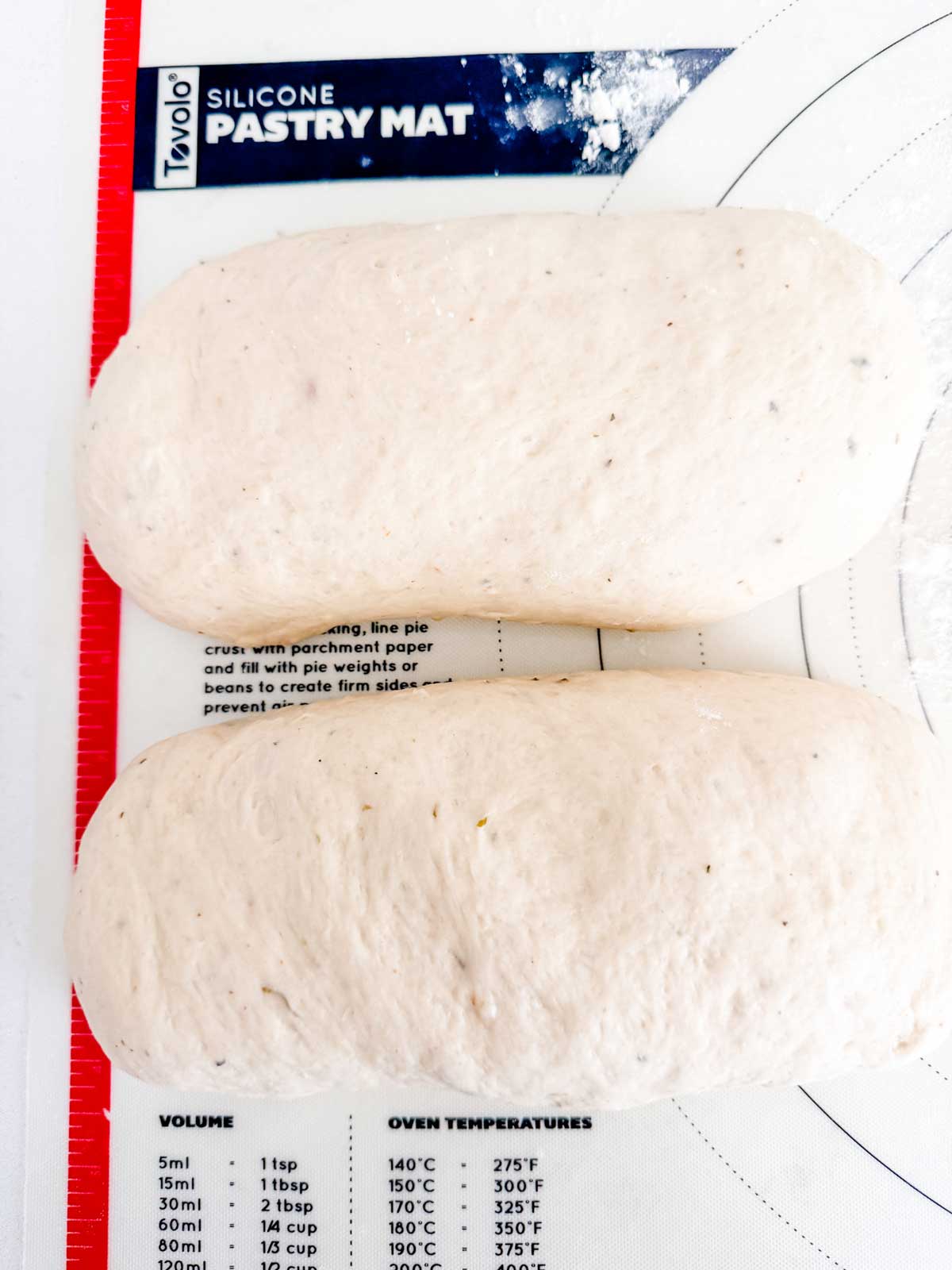 Two oval-shaped, unbaked bread dough loaves rest on a silicone pastry mat with measurement markings and temperature conversion charts visible underneath.