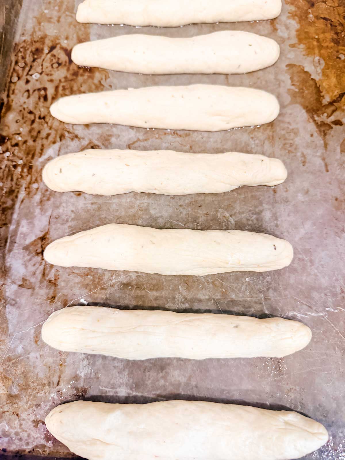 Seven unbaked breadsticks made from dough are evenly spaced on a lightly floured baking sheet, ready for baking.