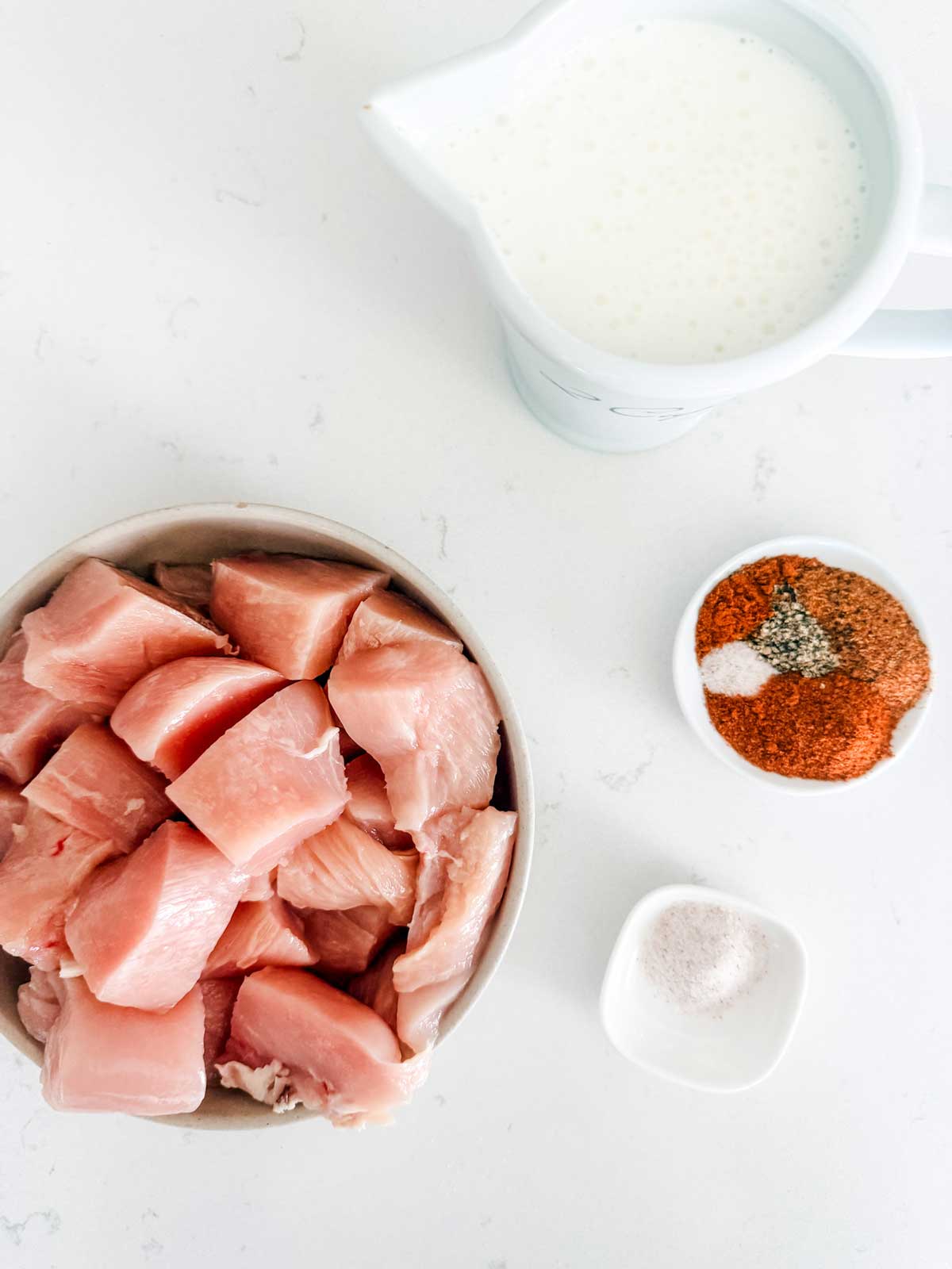 A bowl of raw chicken pieces, a small bowl of mixed spices, a small dish of salt, and a white jug of milk are arranged on a white surface.