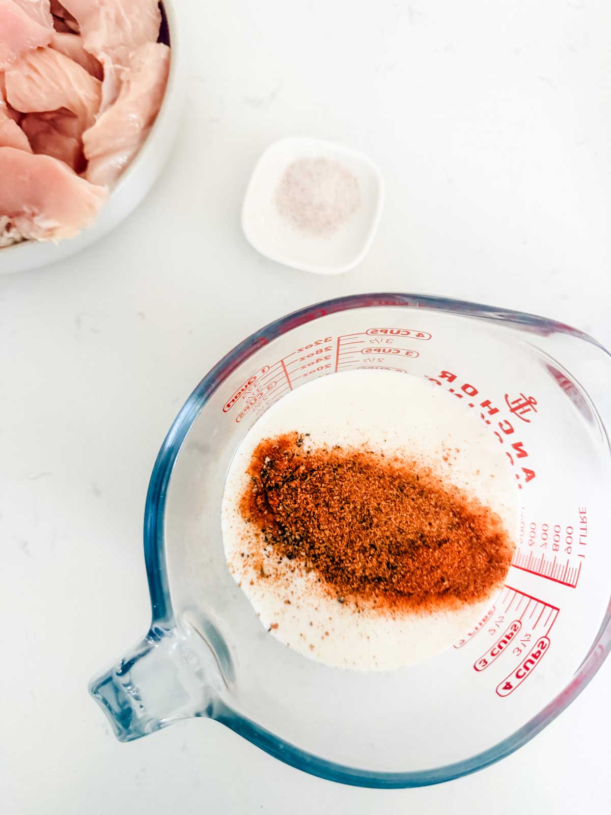 A glass measuring cup with milk and spices, a small dish of salt, and a bowl of raw chicken pieces on a white surface.