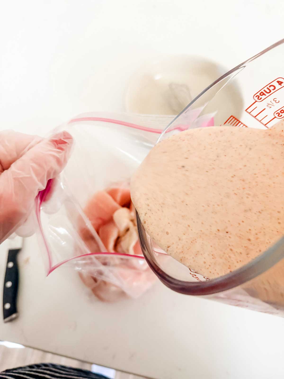 A gloved hand holds a zip-top bag with raw chicken while a creamy marinade is poured from a measuring cup into the bag; a knife and a bowl are on the white counter.
