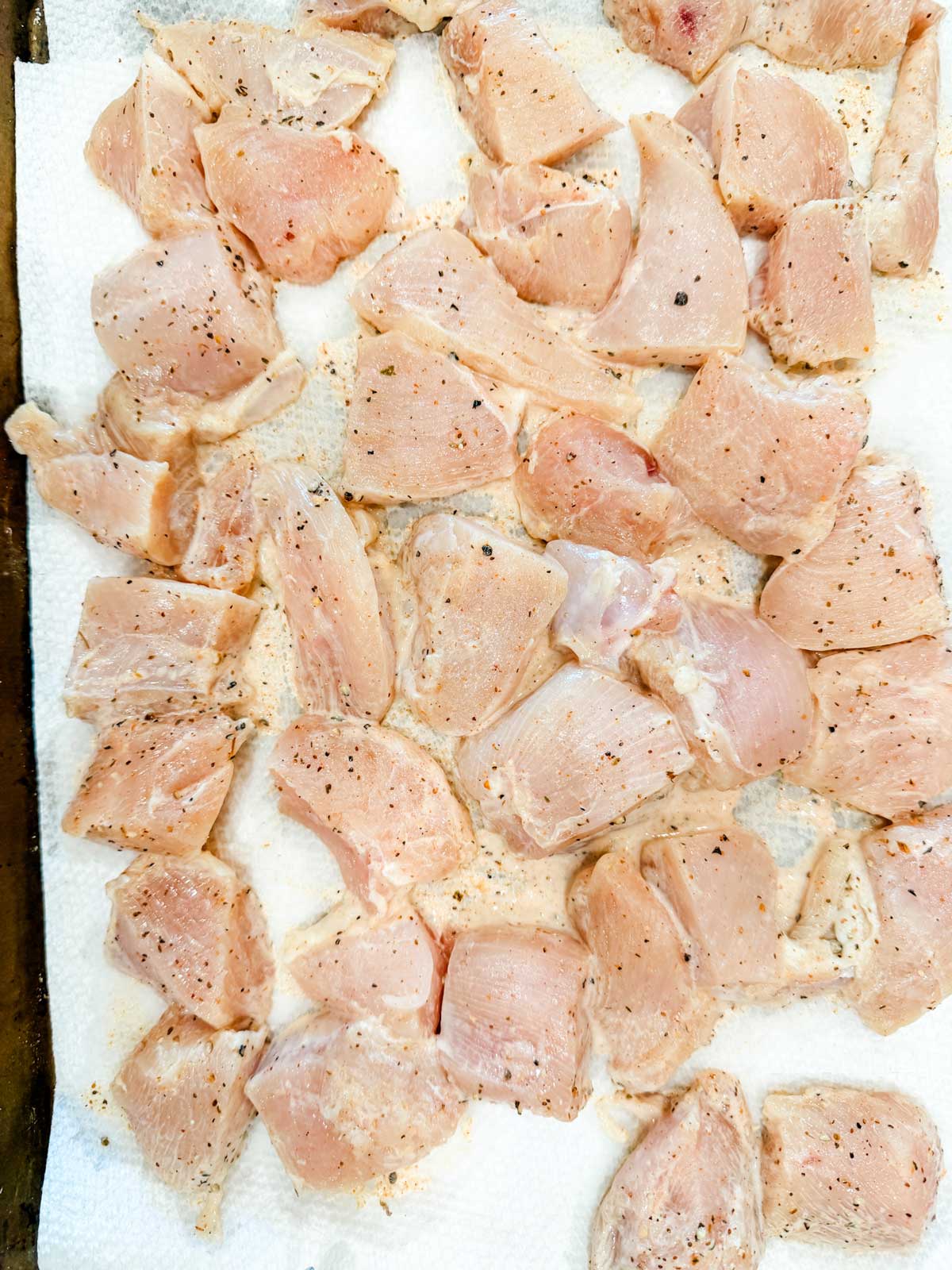 Raw, cubed chicken pieces seasoned with salt and pepper are spread out on a white paper towel, likely to absorb excess moisture before cooking.
