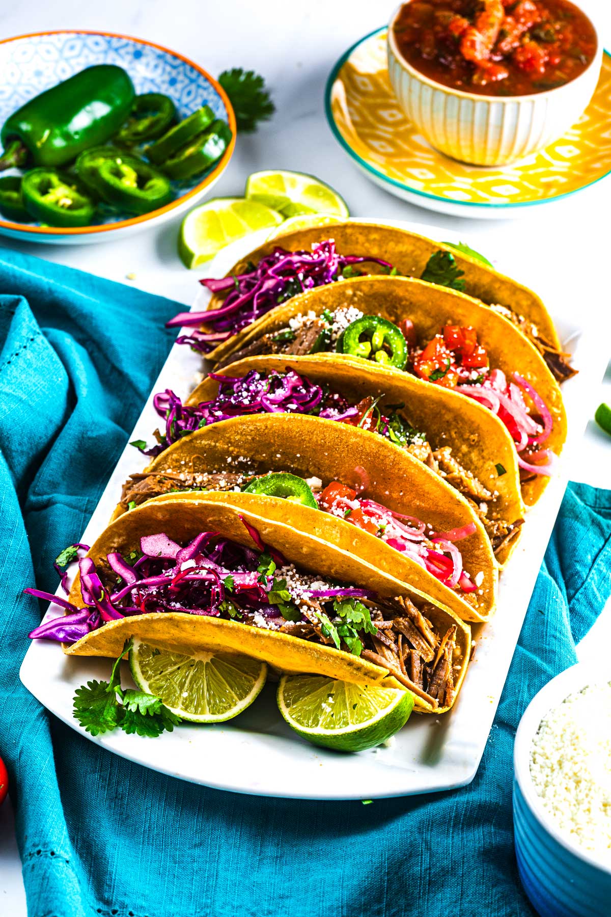 A plate of four beef tacos in crispy shells, topped with shredded red cabbage, sliced jalapeños, cilantro, cheese, and lime wedges, with salsa and sliced jalapeños on the side.