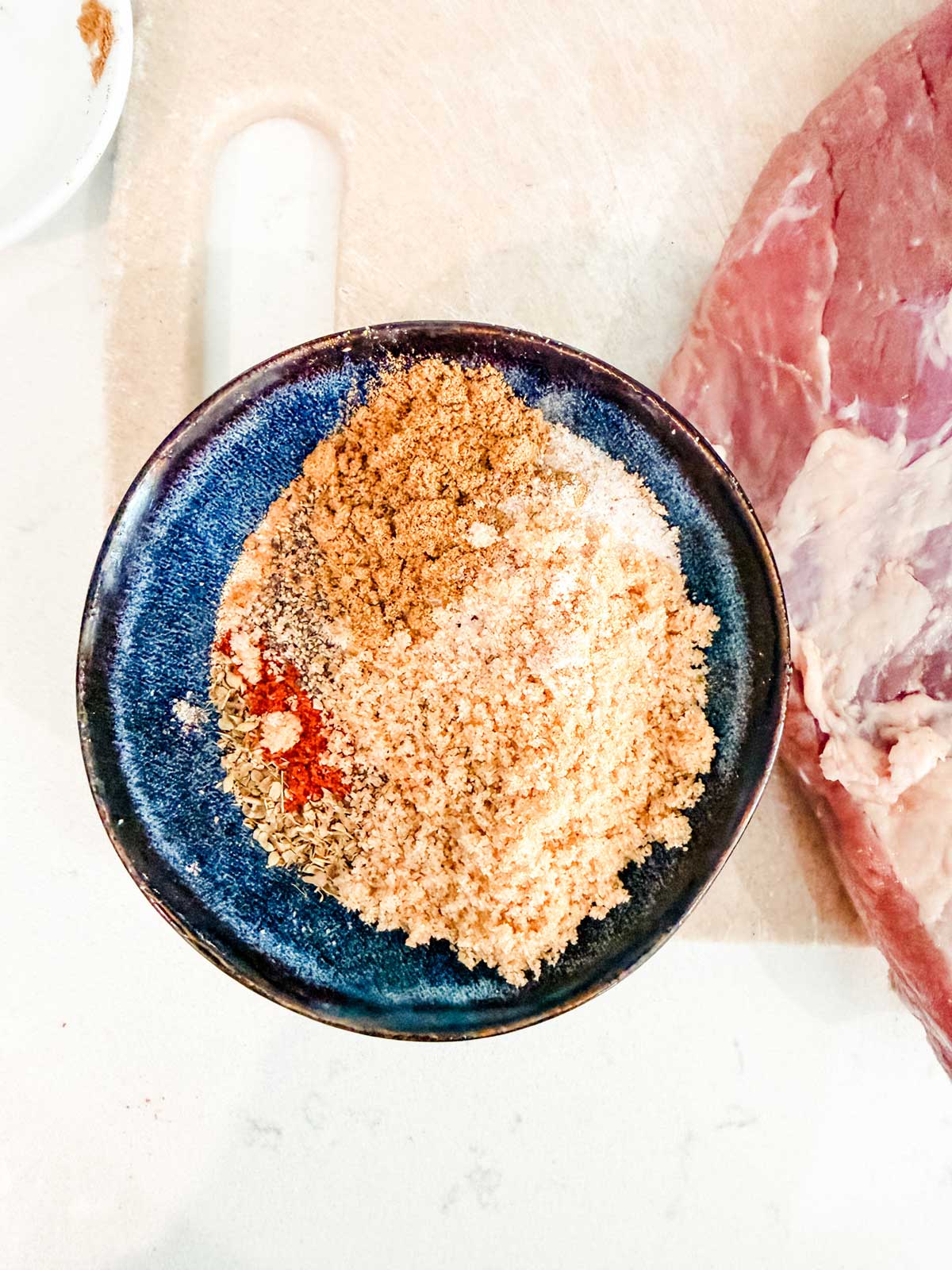 A blue bowl filled with various seasonings and spices sits on a light countertop next to a brisket.