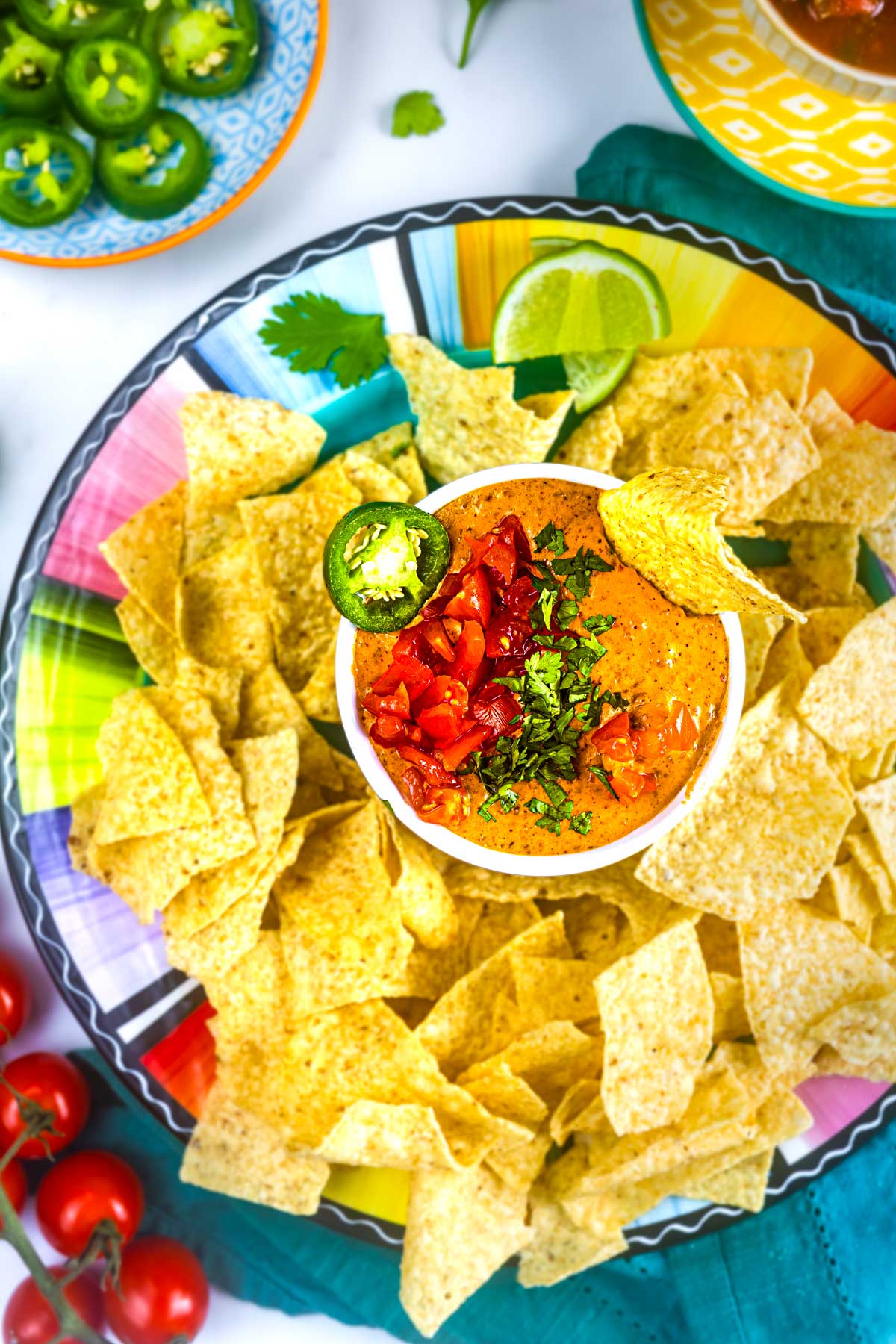 A colorful plate filled with tortilla chips surrounds a bowl of creamy queso dip, topped with chopped tomatoes, cilantro, and a jalapeño slice, with lime wedges nearby. Bright dishes and fresh vegetables are visible around the plate.