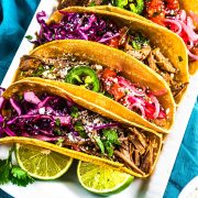 Four tacos filled with shredded meat, purple cabbage, pickled onions, jalapeño slices, tomatoes, cilantro, and crumbled cheese are arranged on a plate, garnished with lime slices, on a blue cloth.