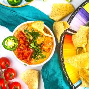 A bowl of cheesy dip topped with tomatoes, cilantro, and a slice of jalapeno sits on a teal cloth, surrounded by tortilla chips, cherry tomatoes, and a colorful plate of more chips.