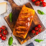 A golden, crusty loaf of bread sits on a wooden cutting board, surrounded by fresh basil leaves and clusters of ripe cherry tomatoes on a light gray surface.