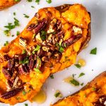 Close-up of baked sweet potato halves topped with chopped pecans, fresh parsley, and a drizzle of honey on a white surface with scattered parsley pieces.