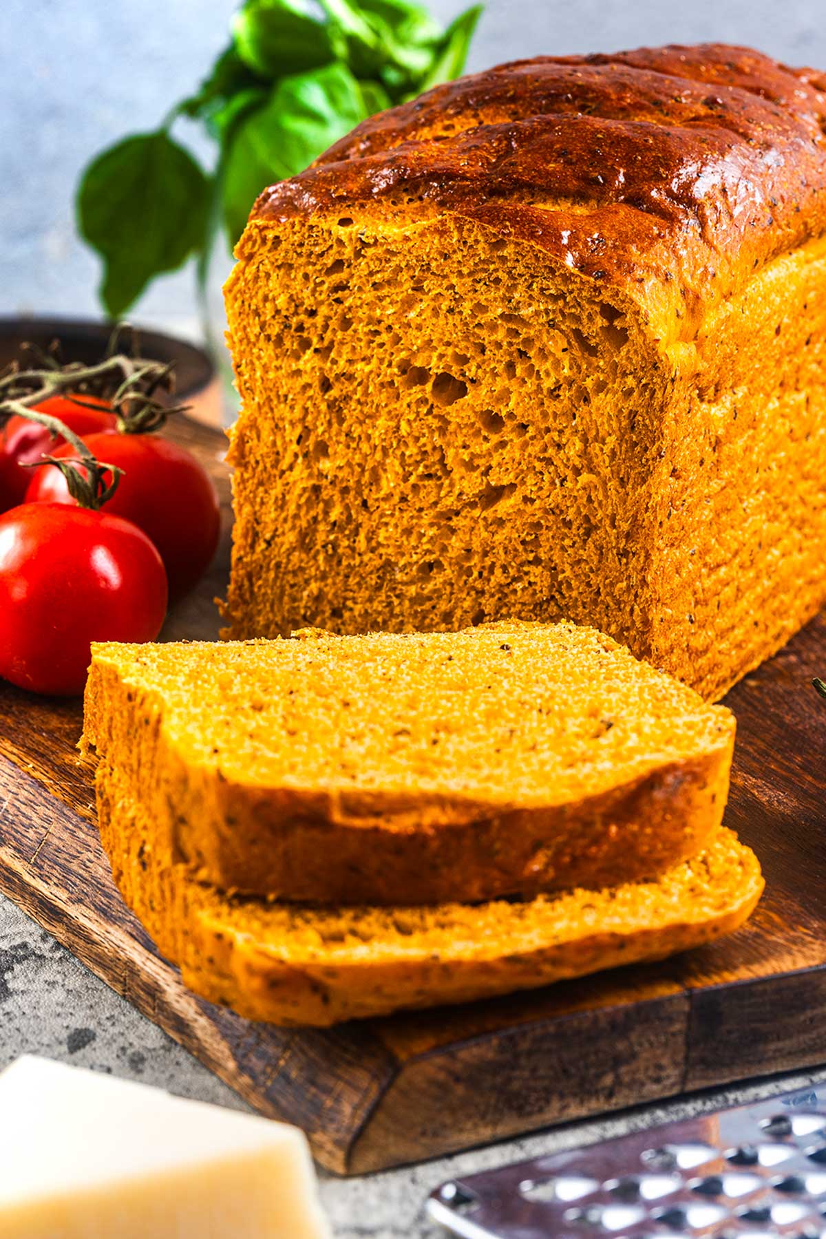 A loaf of golden brown bread sits on a wooden cutting board with two slices cut in front. Fresh tomatoes and basil are in the background, and a cheese grater is visible in the foreground.