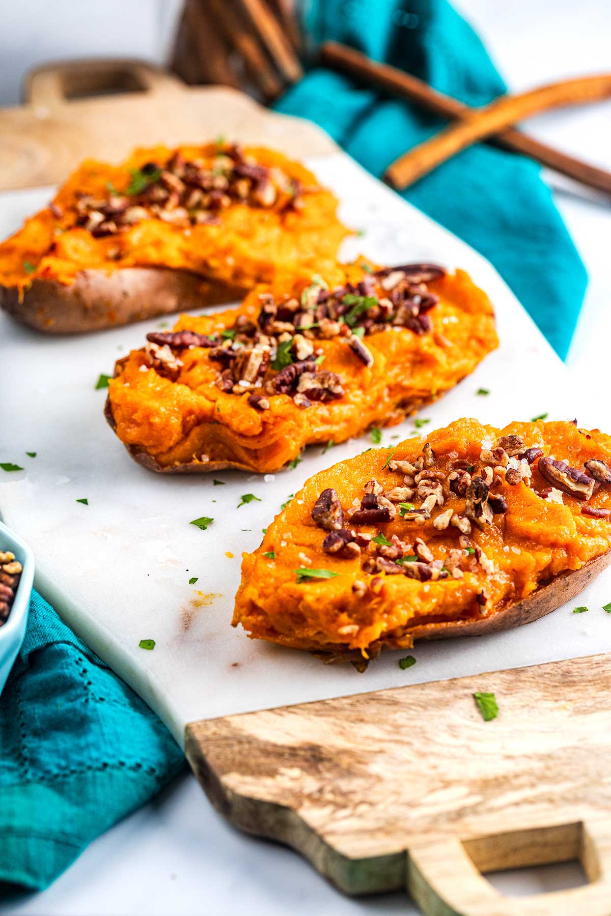 Three twice-baked sweet potato halves topped with chopped pecans and chives are arranged on a white marble board, with a blue napkin and wooden utensils in the background.