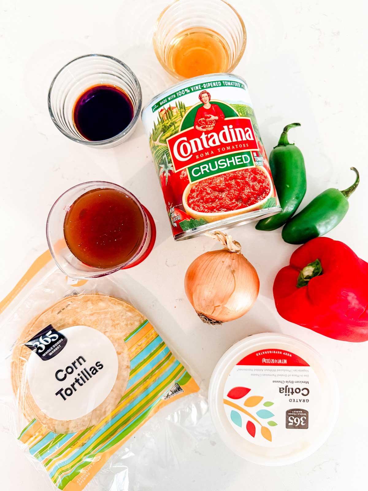 An assortment of ingredients on a white surface, including a can of crushed tomatoes, two jalapeños, a red bell pepper, an onion, corn tortillas, grated cotija cheese, and three small glasses with different liquids.