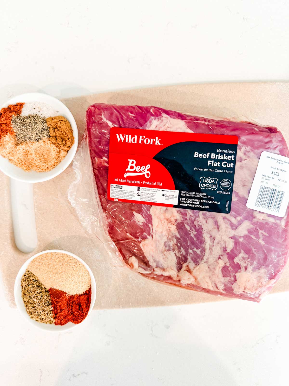 A package of Wild Fork boneless beef brisket flat cut on a cutting board next to two white measuring cups filled with assorted spices and seasonings.
