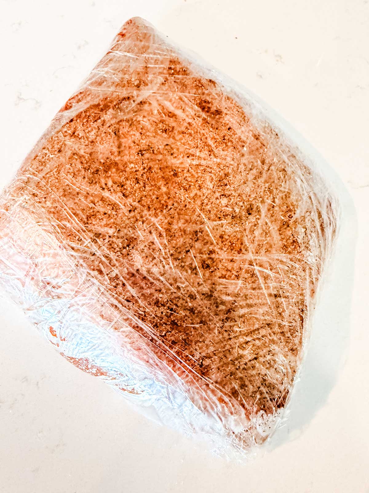 A seasoned brisket wrapped tightly in clear plastic wrap sits on a white surface.