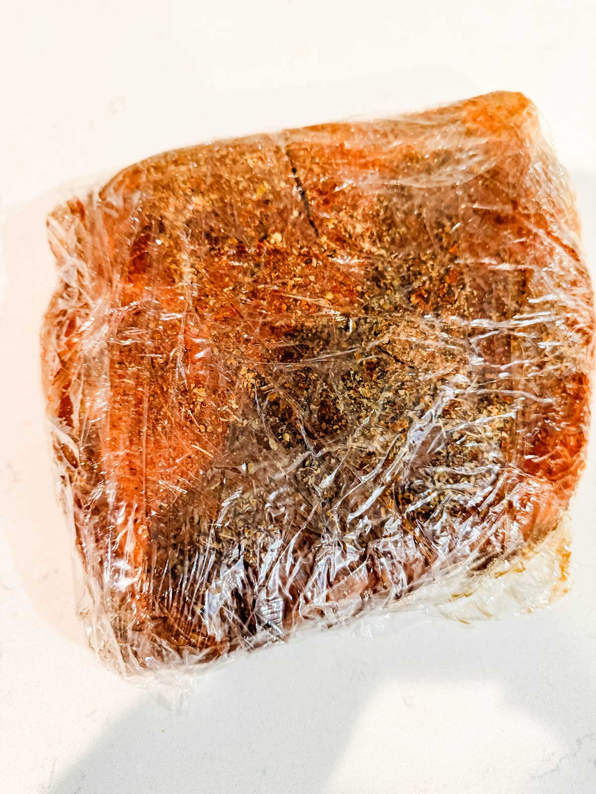 A piece of seasoned meat wrapped tightly in plastic wrap sits on a white surface. The surface of the meat is coated with various spices and herbs.