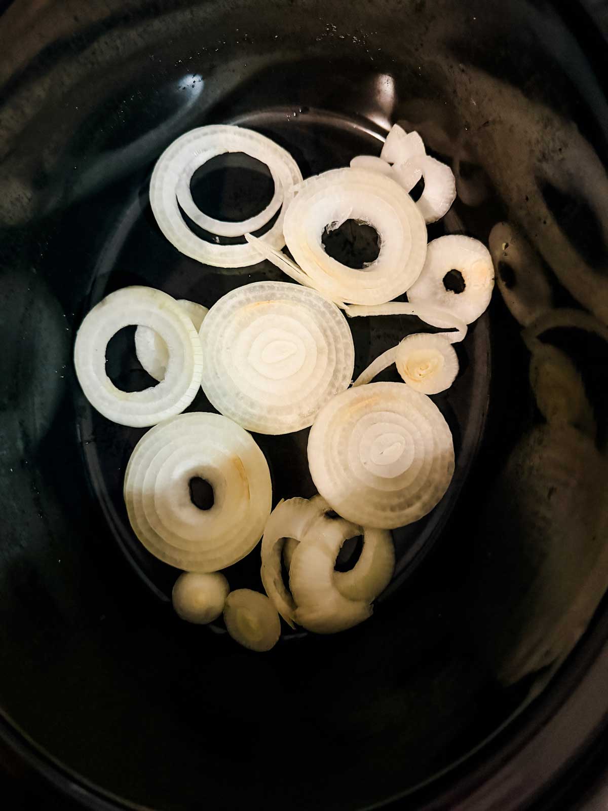 Sliced raw onions arranged in overlapping rings at the bottom of a black slow cooker or pot.