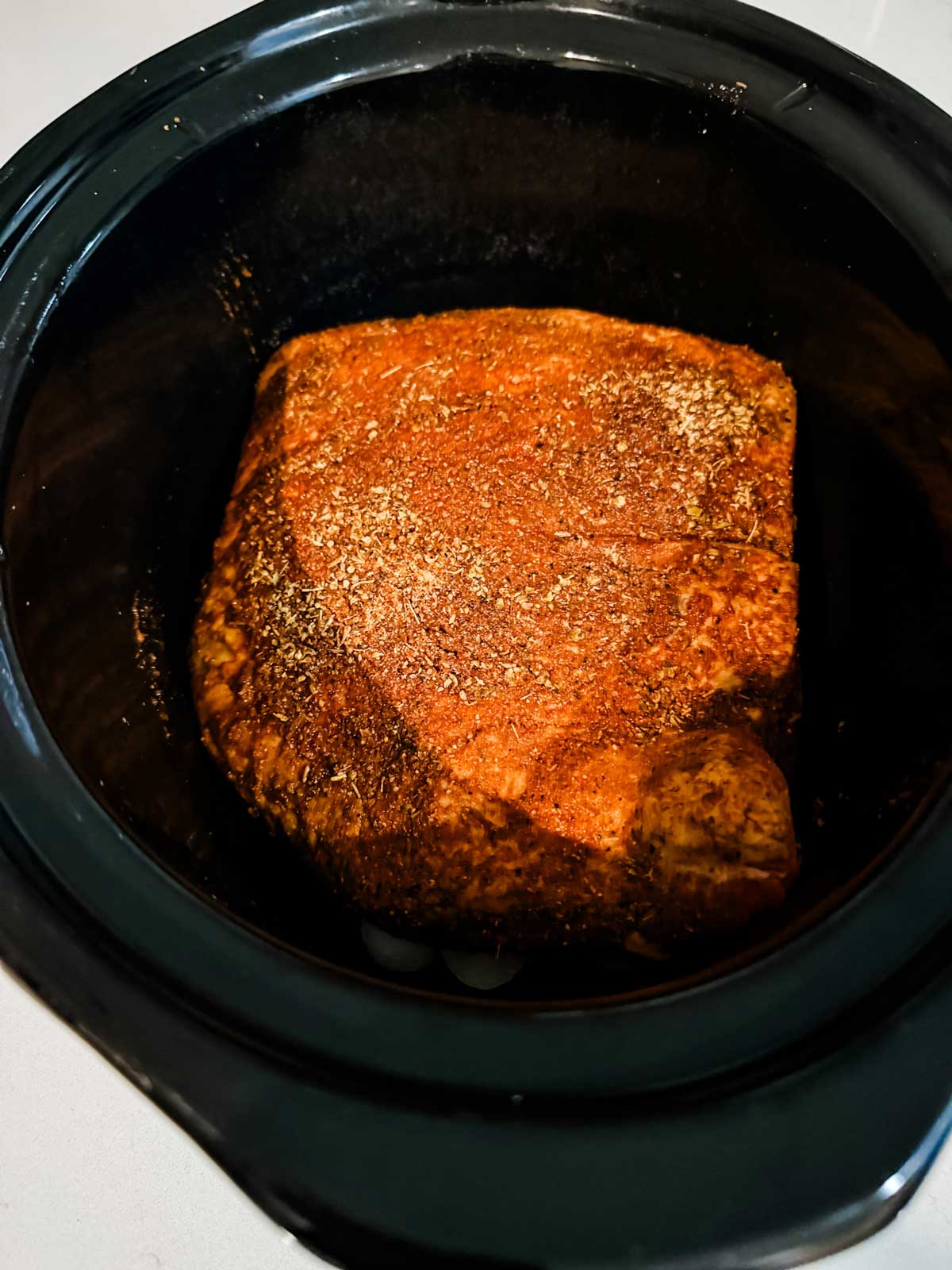 A seasoned piece of meat sits inside a black slow cooker, ready to be cooked. The meat is coated with a dry rub of spices and is uncooked.