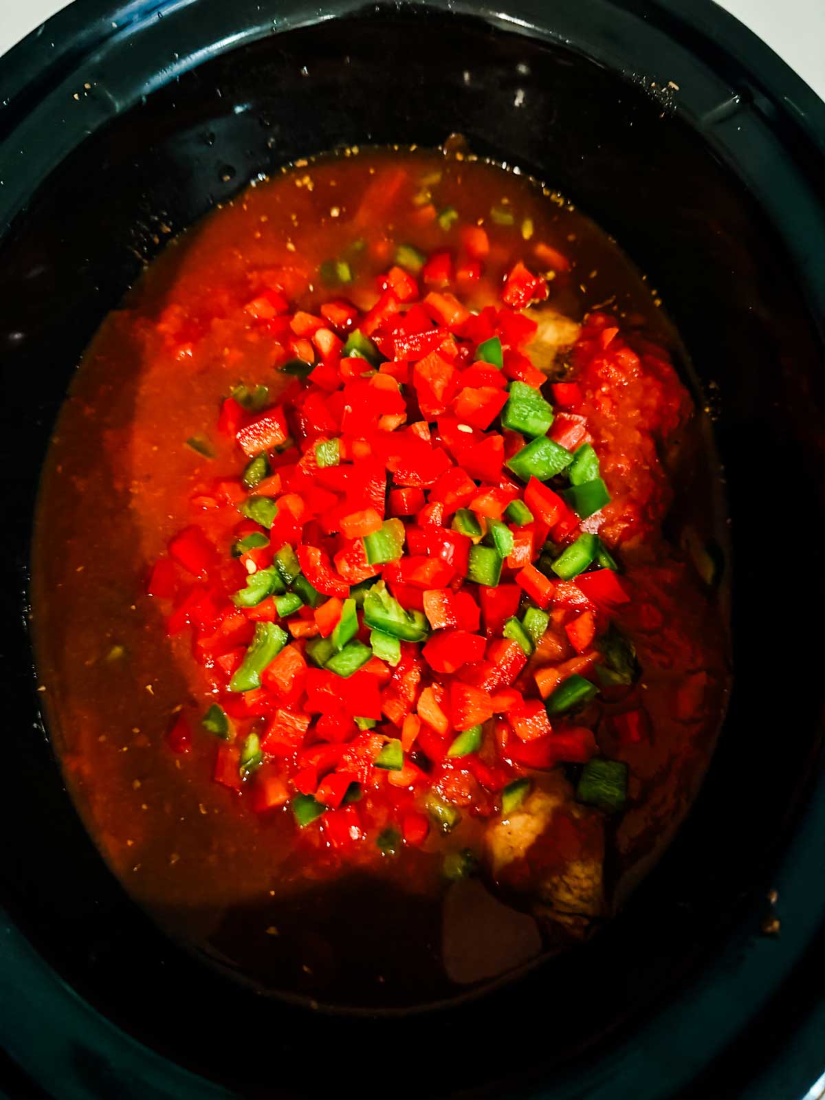 A close-up of a slow cooker filled with a dark, seasoned liquid, pieces of meat, and a topping of finely chopped red and green bell peppers.