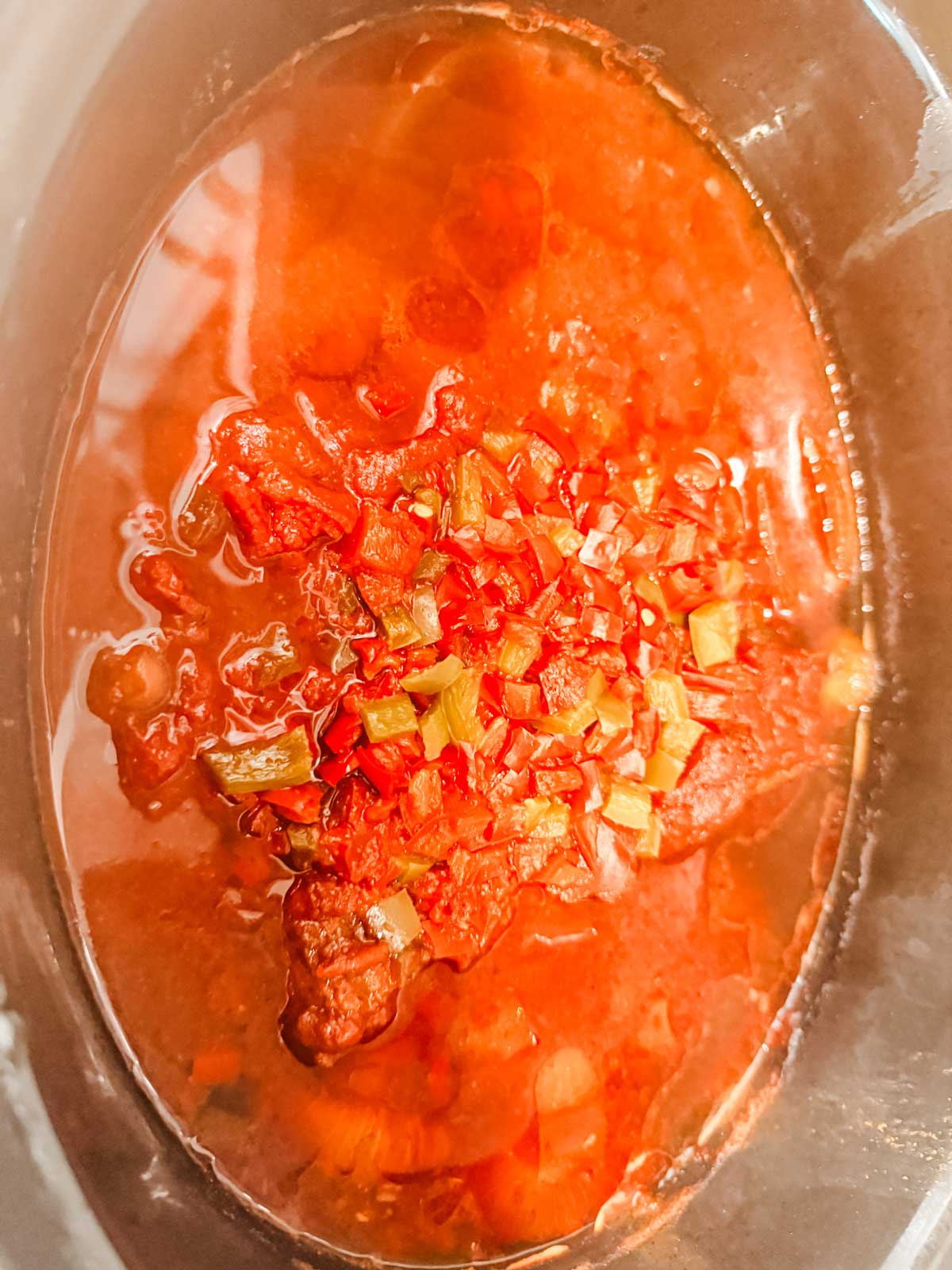 A close-up of a tomato-based stew with chopped red and green peppers in a pot, showing a rich red color and a chunky texture.