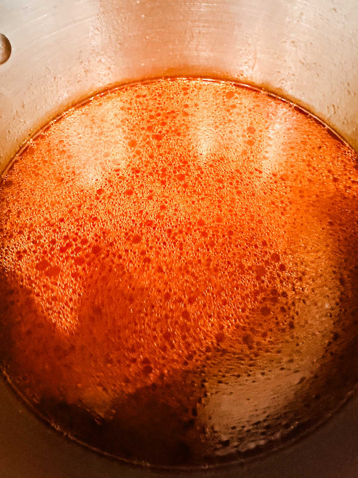 A close-up view of a pot filled with simmering reddish-brown liquid, possibly broth or sauce, with bubbles and steam visible on the surface.