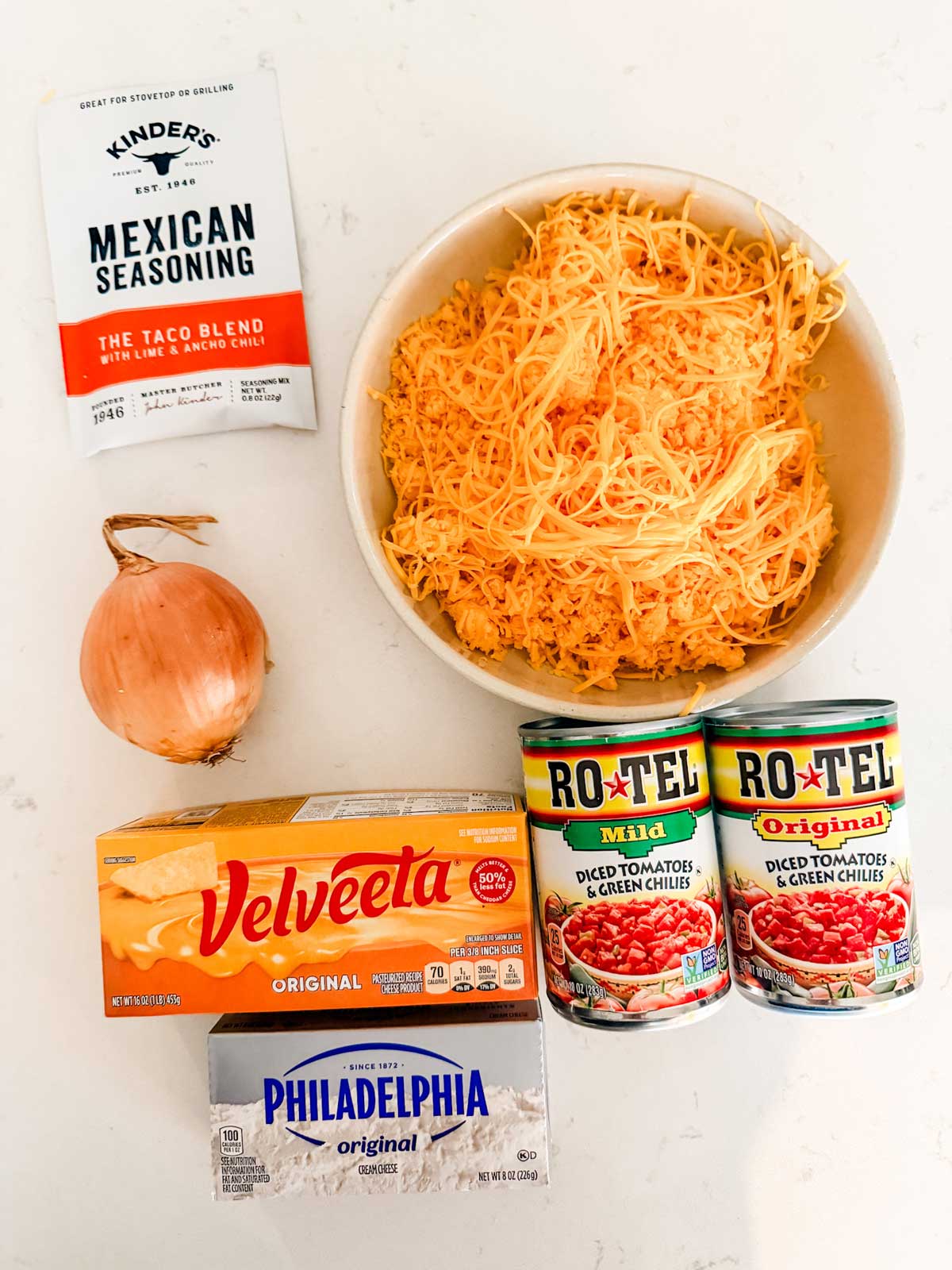 An overhead view of ingredients on a white surface: a packet of Mexican seasoning, a whole onion, a block of Velveeta cheese, a package of Philadelphia cream cheese, shredded cheddar in a bowl, and two cans of Rotel tomatoes with green chilies.