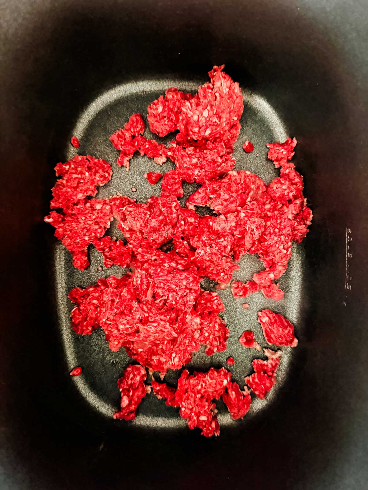 Bright red ground beef scattered in small clumps on the bottom of a black nonstick pan or slow cooker, viewed from above.