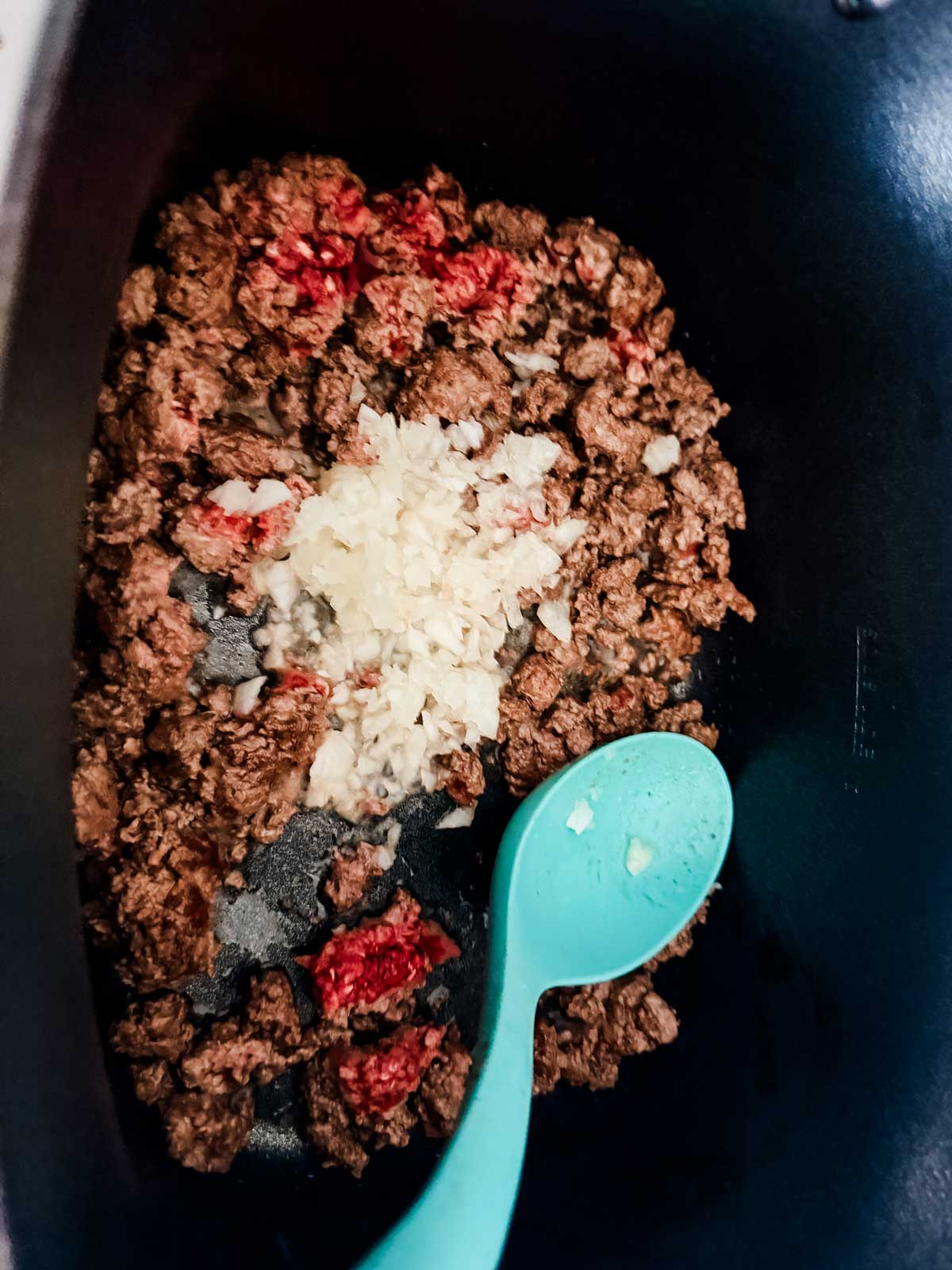 Ground beef is being cooked in a black pot with chopped onions and minced garlic, while a turquoise cooking spoon rests nearby.