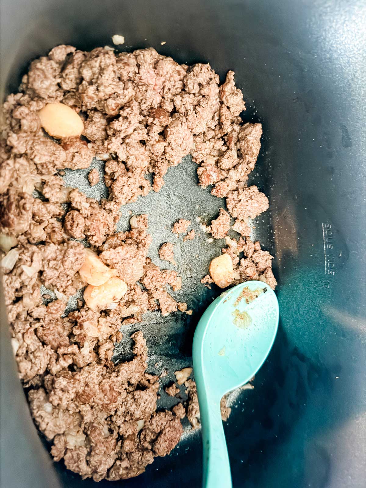 Cooked ground beef and a few chunks of onion in a black pot, with a teal spoon resting inside and some bits of food stuck to the bottom.