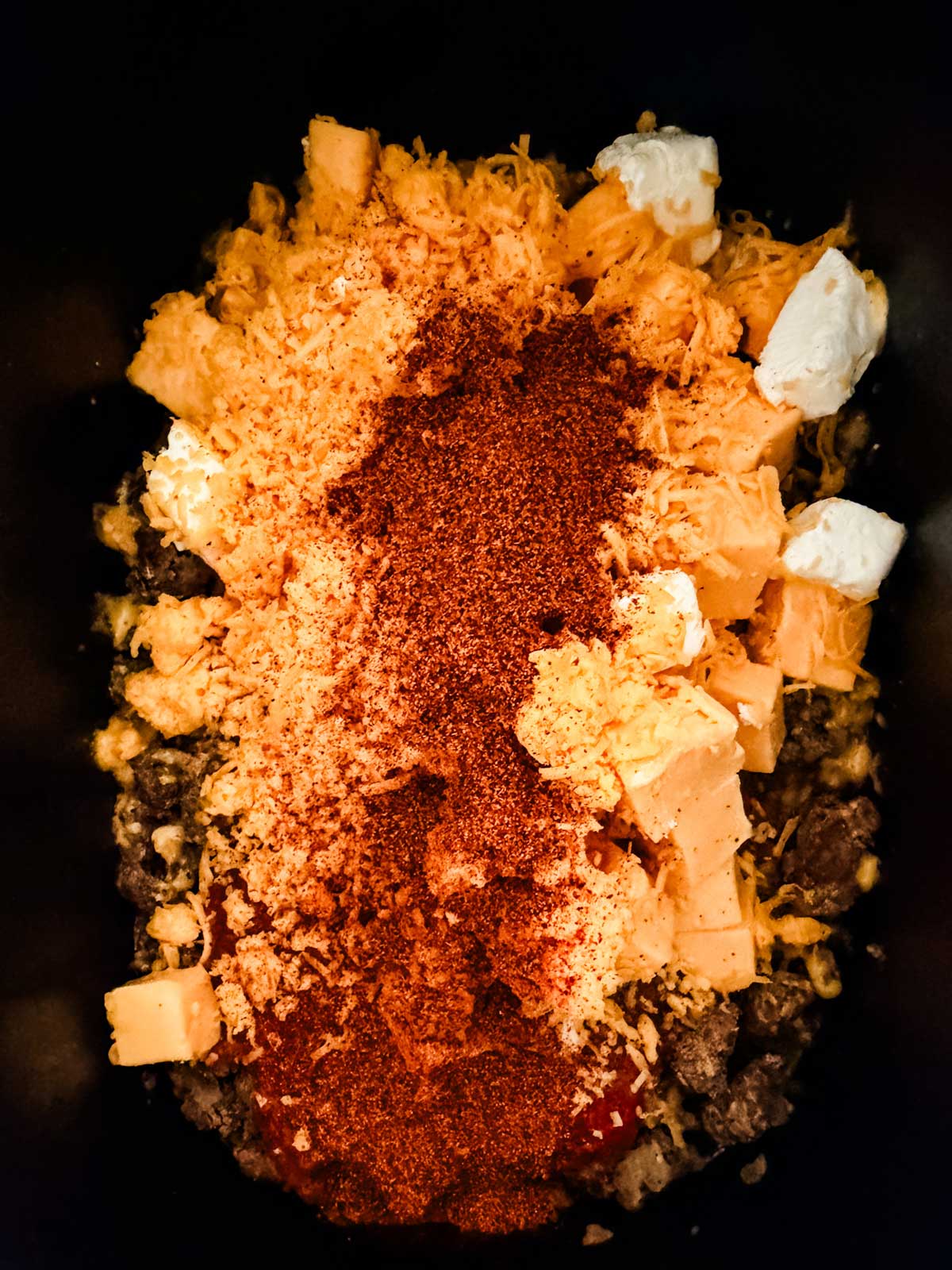 A close-up view of taco seasoning, shredded cheese, cream cheese, and cubed cheese on top of cooked ground beef in a black slow cooker, ready to be mixed.
