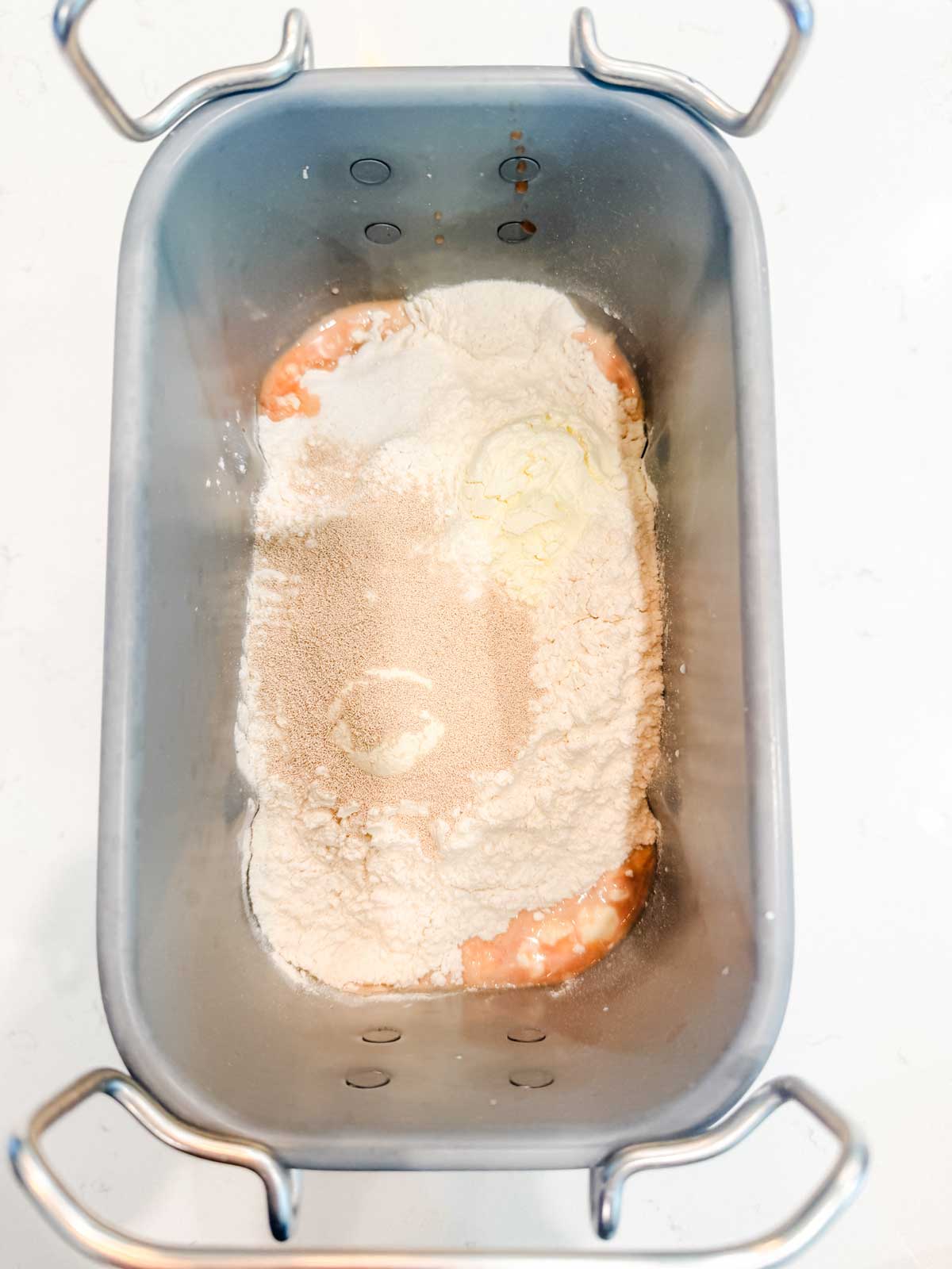 A bread machine pan containing unmixed ingredients, including flour, yeast, and wet ingredients, ready to be mixed and baked.