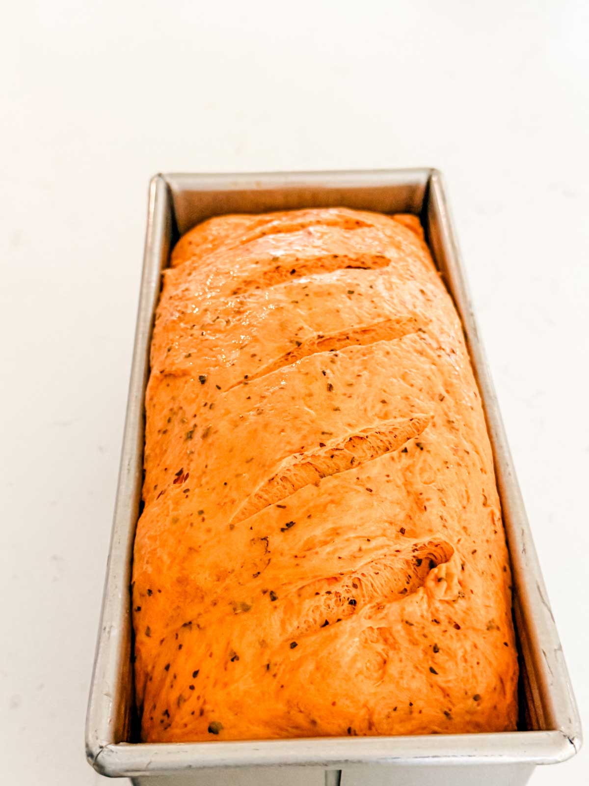 A loaf of unbaked bread dough with herbs, scored on top, sits in a metal loaf pan on a white surface.
