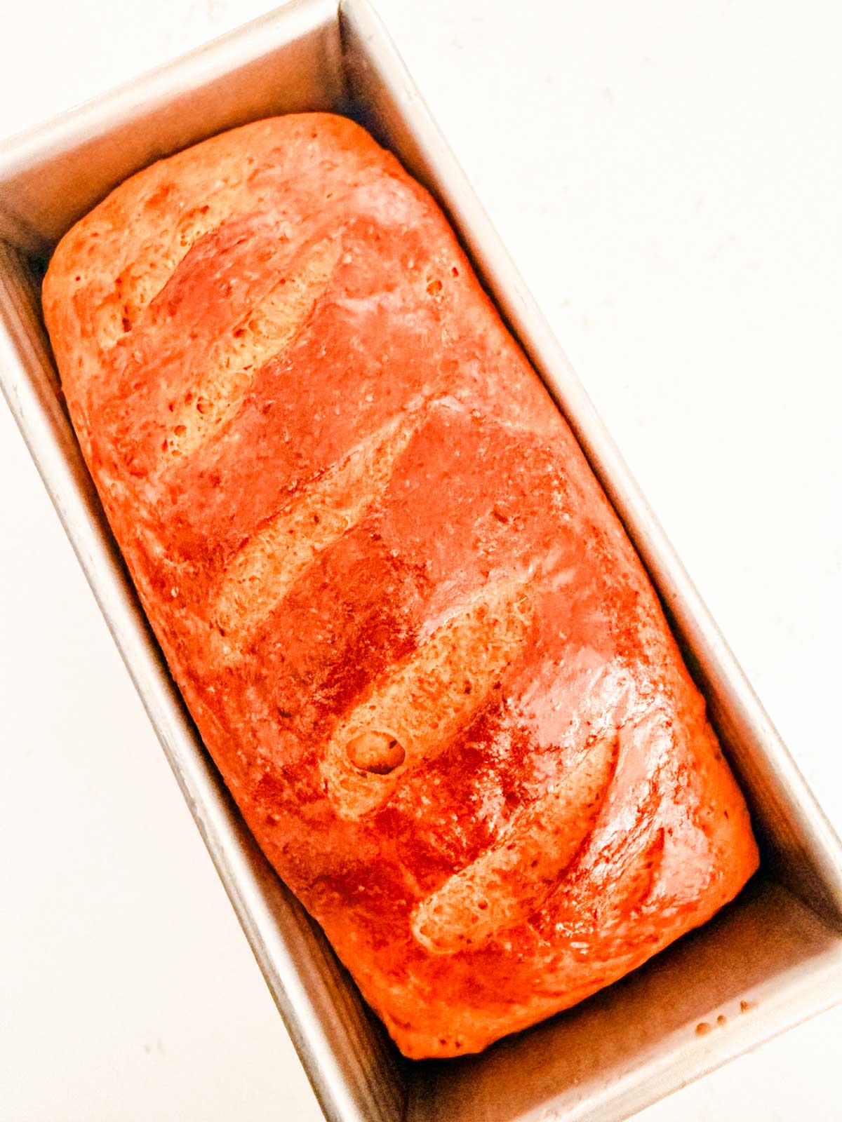 A freshly baked loaf of bread with a golden brown crust sits in a rectangular metal baking pan. The loaf has diagonal slashes on the top and a shiny finish.
