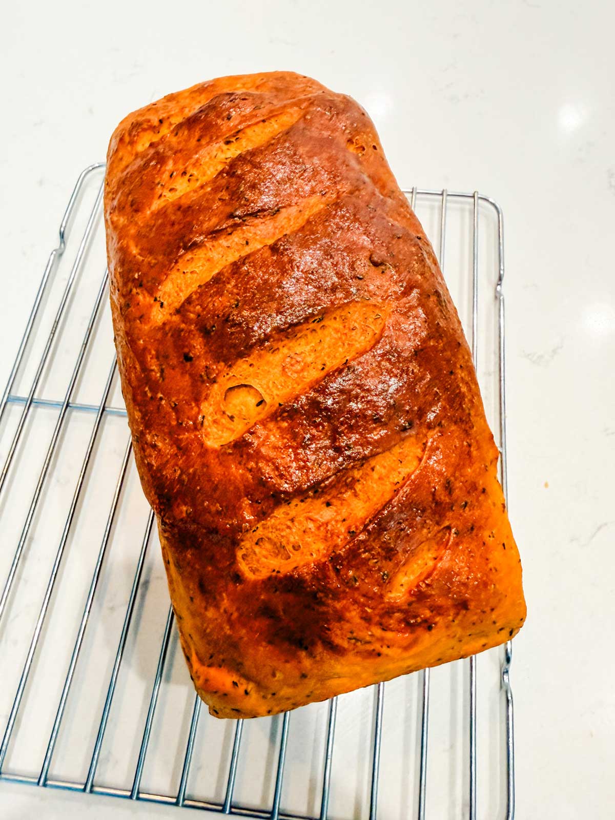 A golden-brown loaf of bread with slashes on top sits on a metal cooling rack, placed on a light-colored countertop.