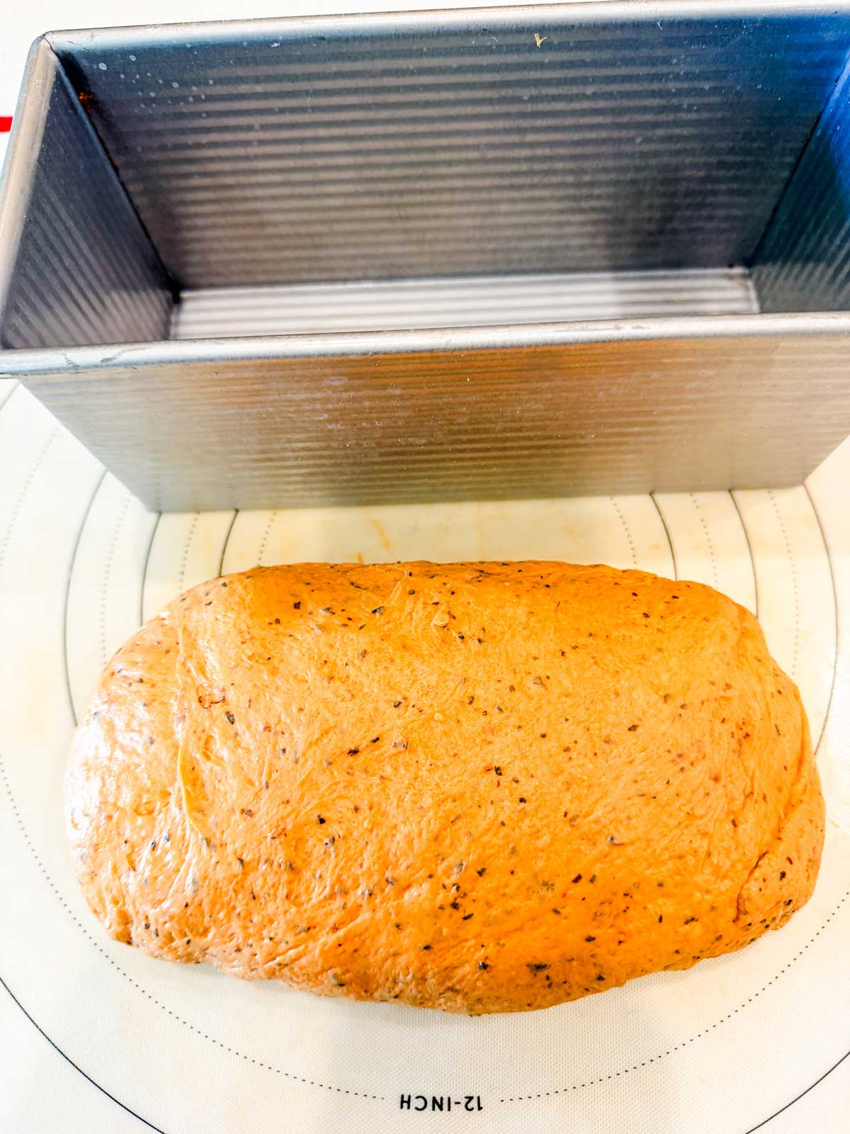 A loaf-shaped ball of brown bread dough with visible seasonings sits on a patterned mat in front of a rectangular metal bread pan.