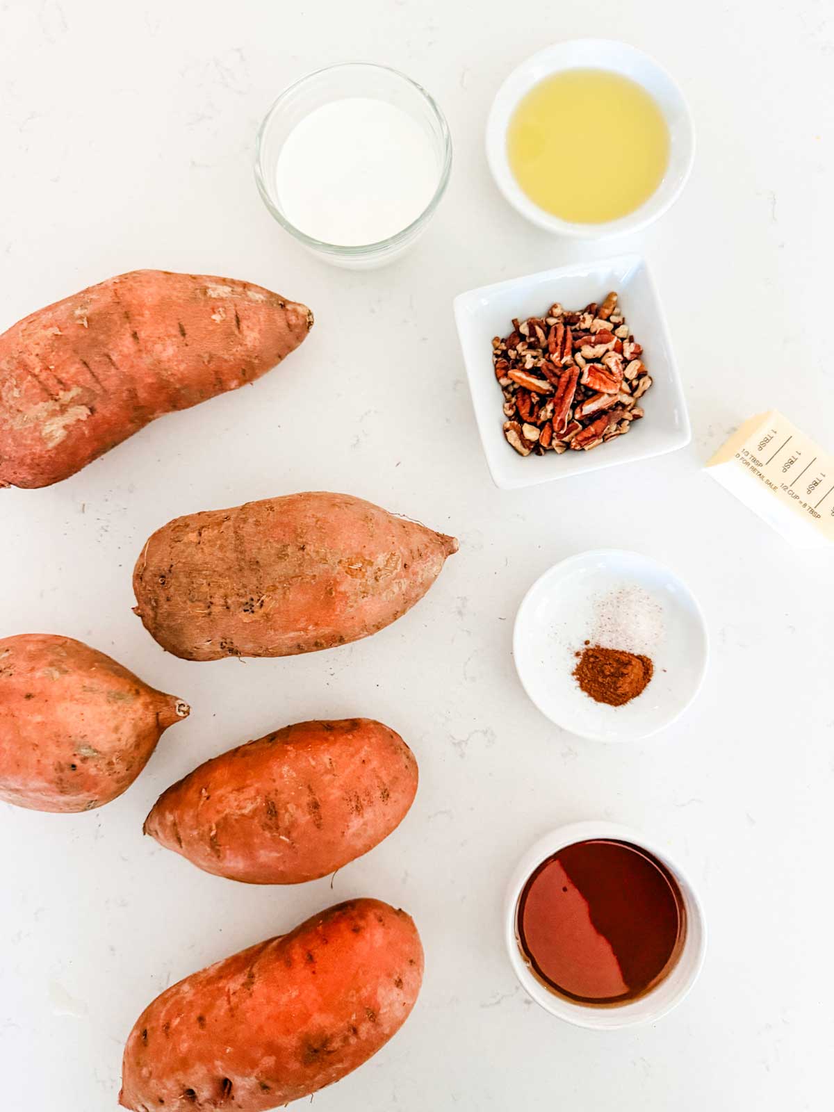Six sweet potatoes, a small dish of chopped pecans, a stick of butter, a bowl of milk, a bowl of oil, a bowl of maple syrup, and a dish with cinnamon and salt arranged on a white surface.