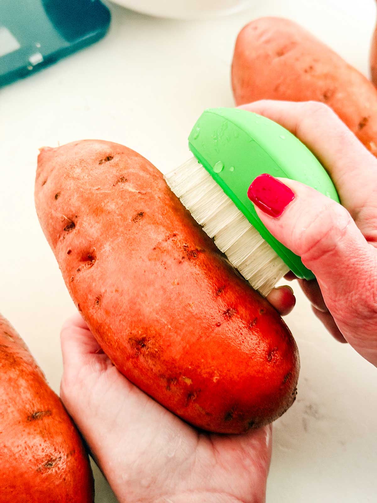A person with red nail polish scrubs a sweet potato with a green and white brush over a white surface. Other sweet potatoes are visible nearby.