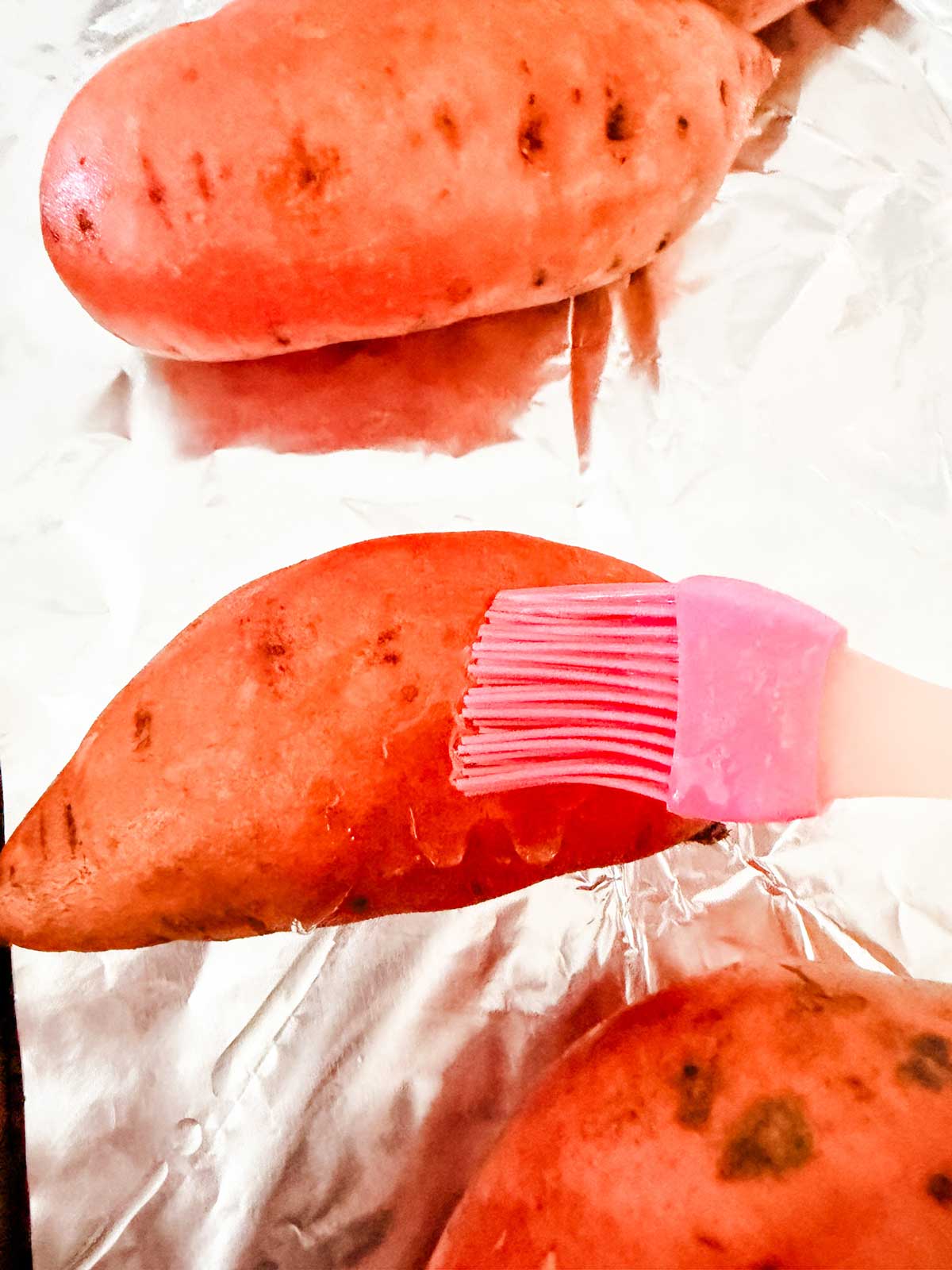 A pink silicone brush is being used to coat whole sweet potatoes with oil on a foil-lined baking sheet.