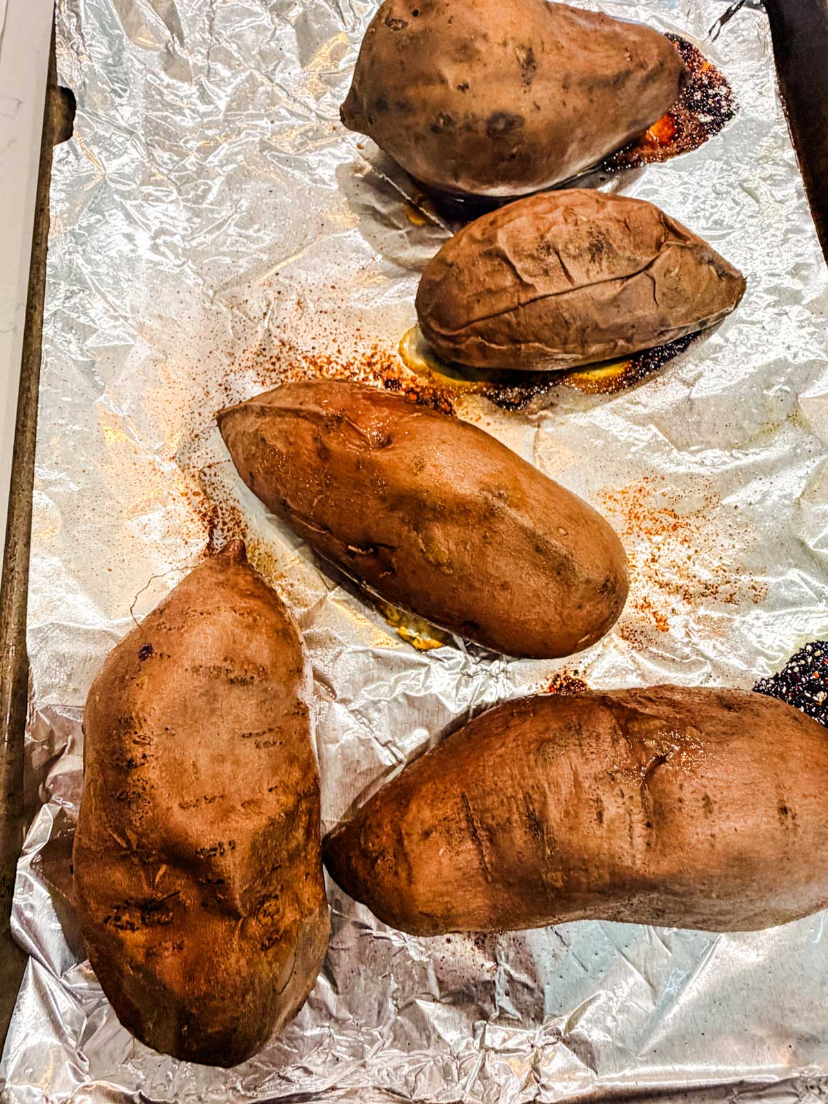 Four baked sweet potatoes and one partially visible sweet potato on a foil-lined baking sheet, showing some caramelized spots and juices.